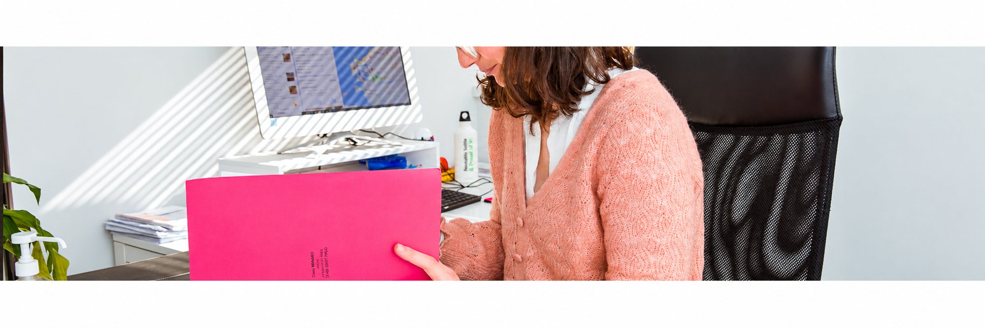 Femme en gilet rose consultant un dossier fuchsia dans un bureau lumineux.